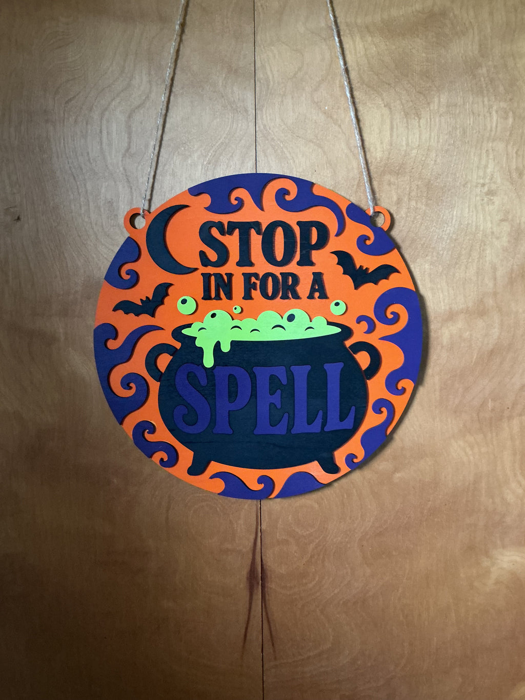 Stop In For A Spell Halloween round door hanger with bubbling cauldron, crescent moon, bats, and filigree in orange and purple layered wood
