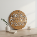 Welcome Floral Garland circular laser engraved wood sign leaning against white wall with neutral home decor