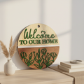 Welcome To Our Home Garden Bloom circular laser engraved wood sign leaning against white wall with neutral home decor